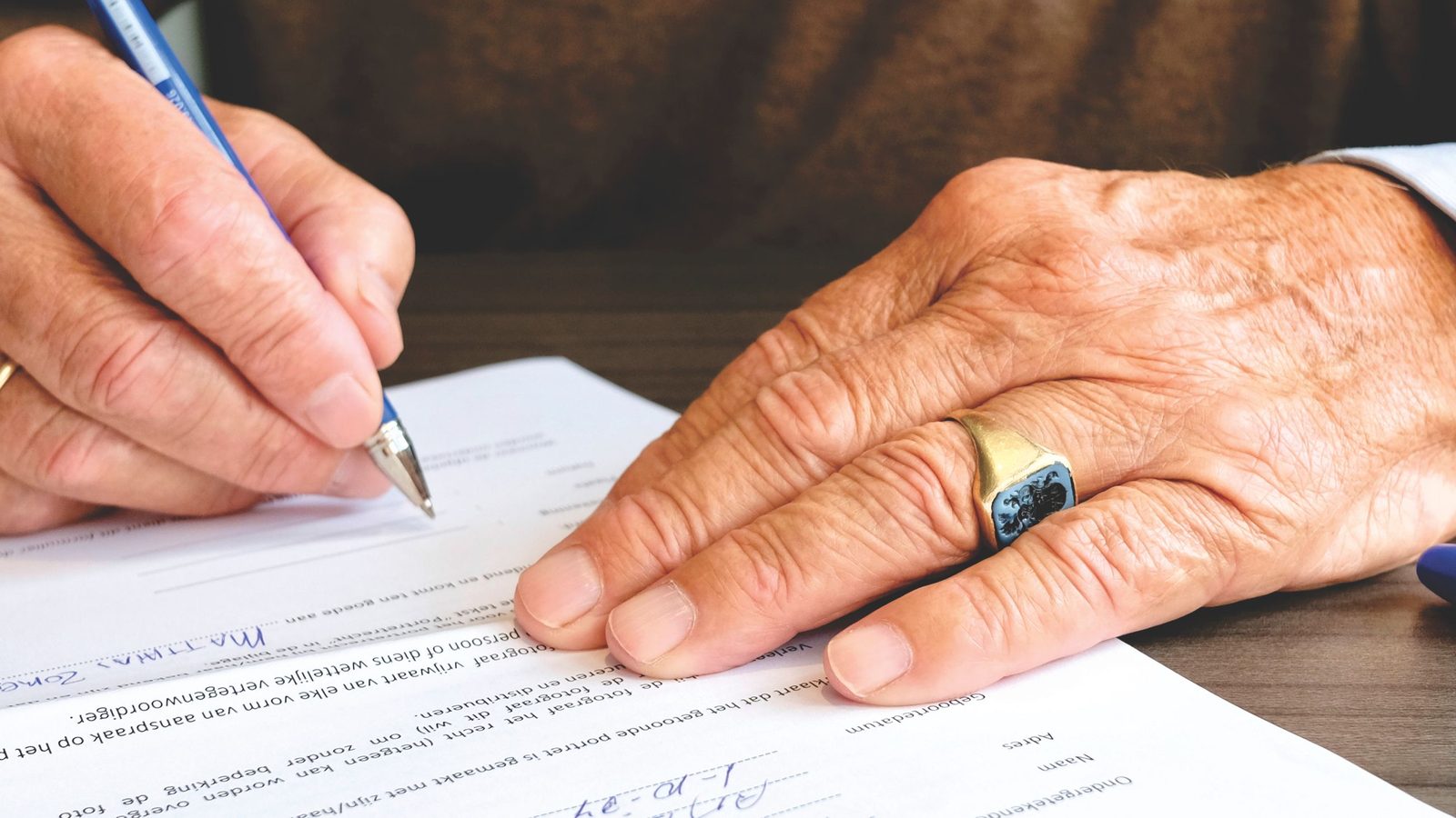 Close-up of an older investor's hands signing an IRA contribution form with a ballpoint pen
