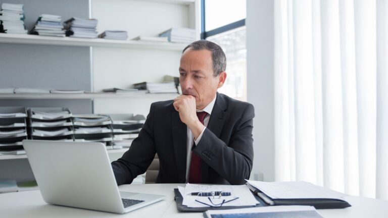 Mature professional in a suit reviewing documents and a laptop at his desk, comparing self-directed IRA fees